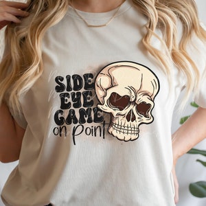 May include: A white t-shirt with a black skull graphic and the text "Side Eye Game On Point".