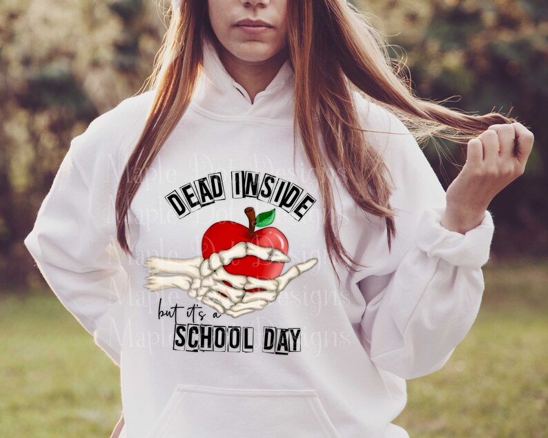 May include: White hoodie with a graphic of a skeleton hand holding a red apple with the text "Dead Inside but it's a School Day" printed on it.