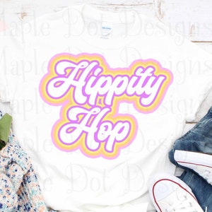Może przedstawiać: Biała koszulka z różowym, żółtym i pomarańczowym nadrukiem z napisem "Hippity Hop" w retro czcionce.