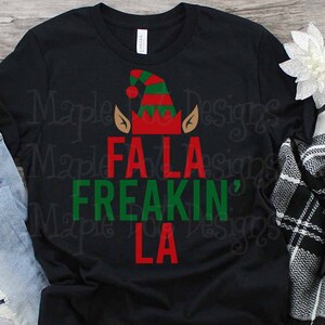 以下が含まれることがあります： 黒いTシャツに、赤と緑のエルフの帽子と「Fa La Freakin' La」という文字がプリントされています。