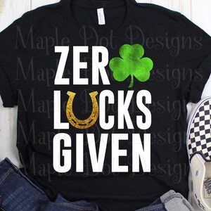 May include: Black t-shirt with white text that reads "ZERO LUCKS GIVEN". A gold horseshoe and a green shamrock are above the text.