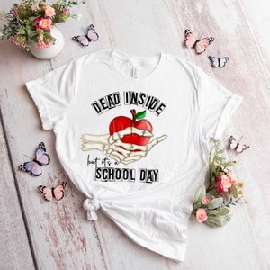 May include: White t-shirt with a black and white graphic of a skeleton hand holding a red apple. The text "Dead Inside but it's a School Day" is printed on the shirt.