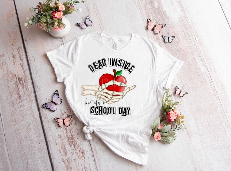 May include: White t-shirt with a black and white graphic of a skeleton hand holding a red apple. The text "Dead Inside but it's a School Day" is printed on the shirt.