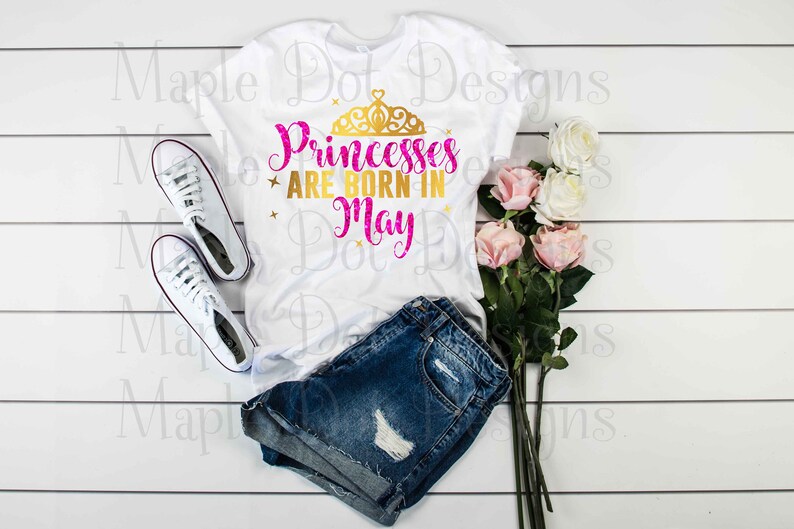 May include: White t-shirt with the text "Princesses are born in May" in pink and gold glitter lettering with a gold crown above the text. The shirt is lying on a white wooden surface with a pair of white trainers and a bouquet of pink roses.