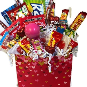 May include: A red gift box filled with an assortment of candies, including Starburst, Twizzlers, and other treats. The box is decorated with heart patterns and filled with white paper shreds. A pink lollipop and heart-shaped decorations are also visible.