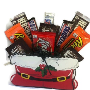 May include: A festive candy arrangement in a Santa boot-shaped container. The red boot features a white trim and gold buckle accents. Assorted chocolate bars and candies, including Reese's, Hershey's, and Snickers, are arranged inside.