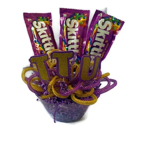 May include: Purple and gold gift basket with three Skittles candy bars and the word "IT'S" in gold glitter letters.