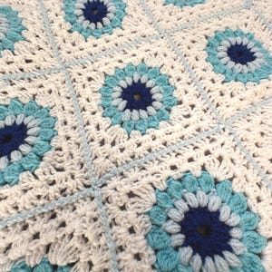 May include: A crochet blanket featuring a repeating square pattern. Each square contains a circular motif with a dark blue centre, surrounded by concentric rings of light blue and turquoise yarn, all set against a white background.