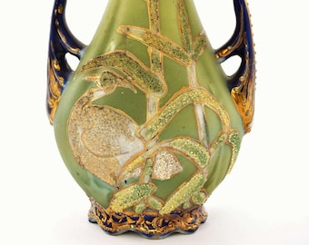 May include: A decorative ceramic vase with a light green body, adorned with gold and green plant motifs. The vase features dark blue handles and trim with gold accents. The design includes a bird and plant life. The vase is approximately 10 inches tall.