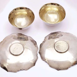 Peut inclure: Quatre pièces de service en argent. Deux petits bols ronds avec des intérieurs dorés sont au-dessus de deux grandes assiettes de forme irrégulière. Les assiettes ont un petit centre rond argenté. Les pièces sont disposées sur une surface blanche.