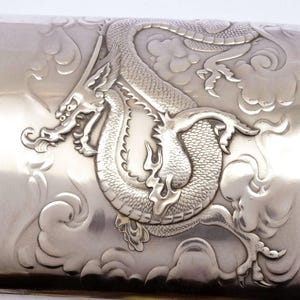 May include: A polished silver-toned metal flask featuring a detailed embossed design of a coiled dragon surrounded by stylized clouds. The metallic surface has a reflective finish, highlighting the intricate texture and relief of the dragon motif.