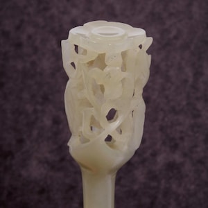 May include: A carved, off-white jade object with a floral design at the top. The jade is intricately carved with open spaces and a smooth, polished finish. The stem is long and slender, leading to the detailed floral top.