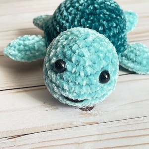 May include: A handmade crochet turtle in shades of teal and turquoise. The turtle has a round head with black safety eyes and a small smile. The plush toy is made of soft, textured yarn and is a cute, whimsical design.