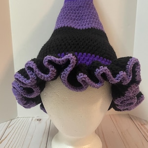 May include: A crocheted witch hat featuring a pointed crown with alternating horizontal stripes of purple and black yarn. The wide, circular brim has a ruffled edge finished with a purple yarn border, creating a distinct wavy texture throughout.