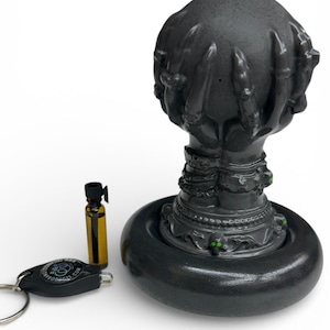 May include: A dark gray decorative object with a hand-shaped top, a round base, and a detailed pedestal. A small bottle and a keychain are next to it. The object has green accents.