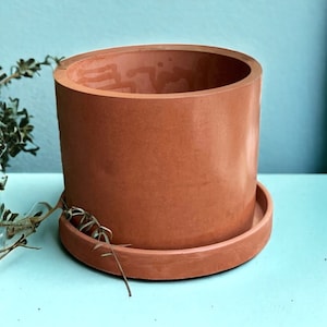 May include: A terracotta colored ceramic planter with a matching saucer. The planter is cylindrical in shape and has a smooth, slightly textured surface.