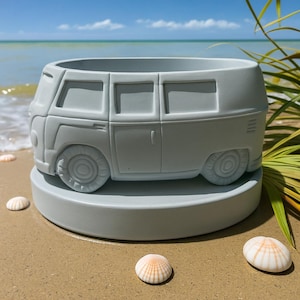 May include: A light gray planter shaped like a vintage van, with detailed wheels and windows. The planter sits on a round base, with seashells scattered around it on a sandy surface. The background features a blue sky and ocean.