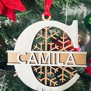 May include: White wooden Christmas ornament shaped like the letter C with a snowflake design and the name "CAMILA" written across the center.