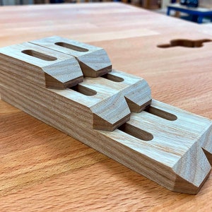 May include: A stack of wooden woodworking clamps with a natural wood grain finish. Each clamp has a rectangular body with a slot and angled edges. The clamps are arranged in a stepped configuration, showcasing their design.
