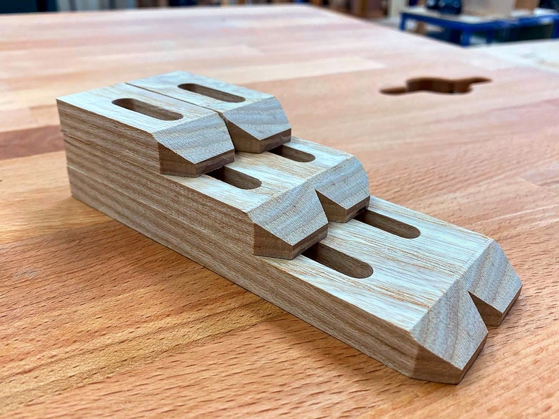 May include: A stack of wooden woodworking clamps with a natural wood grain finish. Each clamp has a rectangular body with a slot and angled edges. The clamps are arranged in a stepped configuration, showcasing their design.