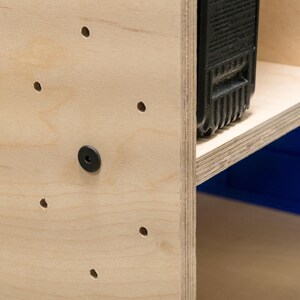 Puede incluir: Primer plano de una estanter&iacute;a de madera clara con una bater&iacute;a de herramienta el&eacute;ctrica negra en el estante superior. La unidad tiene varios agujeros perforados en el lateral para el montaje.