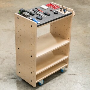 Puede incluir: Un carro rodante de madera con tres estantes y una parte superior de tablero perforado. El carro est&aacute; hecho de madera contrachapada y tiene ruedas negras. El estante superior est&aacute; cubierto de espuma negra y tiene varias herramientas y suministros.