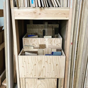 Puede incluir: Un carro de almacenamiento de madera con tres cajones, cada uno lleno de diferentes piezas de madera. El carro est&aacute; sobre ruedas y est&aacute; colocado frente a una pared de paneles de madera apilados.