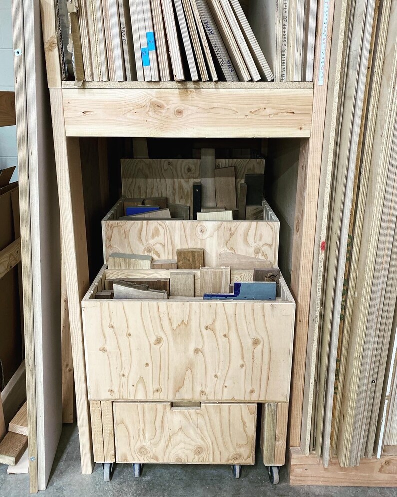 Puede incluir: Un carro de almacenamiento de madera con tres cajones, cada uno lleno de diferentes piezas de madera. El carro est&aacute; sobre ruedas y est&aacute; colocado frente a una pared de paneles de madera apilados.