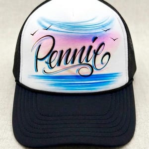 May include: A trucker hat with a white front panel featuring a colorful airbrushed design of a sunset over water, with the name "Pennie" in cursive script. The brim and mesh back are navy blue.