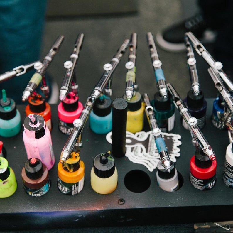 Pu&ograve; includere: Un supporto organizzatore nero che contiene diversi aerografi argentati e vari piccoli flaconi di plastica riempiti con vernici colorate, tra cui tonalit&agrave; rosa, giallo, rosso, blu e verde, disposti a griglia sulla superficie.