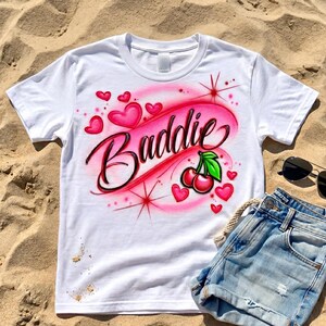 Cherry Airbrush design on a white shirt layed out on the sand. Baddie with black lettering and red with pink background.