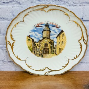 May include: A white ceramic plate with a gold rim and a painted scene of a church in St. Aurelien, Limoges. The plate has a scalloped edge and a raised design.