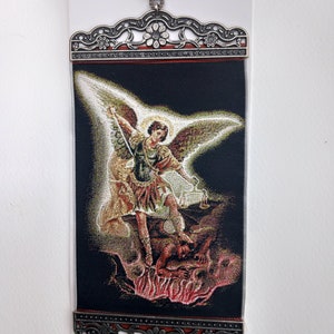 Op de afbeelding: Een wandtapijt met een zilveren frame dat een engel met vleugels afbeeldt, die een zwaard vasthoudt en boven een demon in vlammen staat.