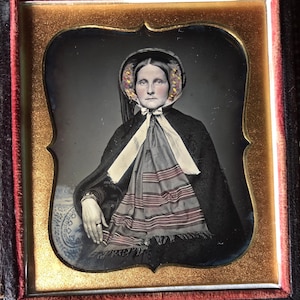 以下が含まれることがあります： 黒いケープとボンネットを身につけた女性のヴィンテージポートレート。 ボンネットにはカラフルな花のアクセントが施されています。 彼女はストライプのスカーフと白いリボンを着用しています。 ポートレートは金と赤で額装されており、歴史的なファッションを紹介しています。