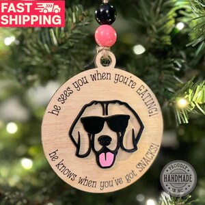 May include: A round wooden bauble with a black and white illustration of a dog wearing sunglasses. The bauble has the text "He sees you when you're eating! He knows when you've got snacks!" printed on it.