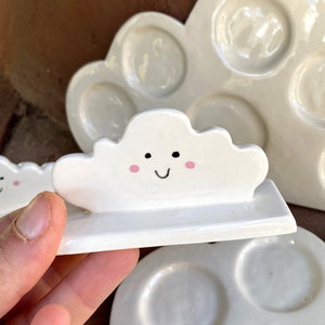 May include: Three white ceramic paint palettes shaped like clouds. The smallest palette has a smiling cloud face with pink cheeks.