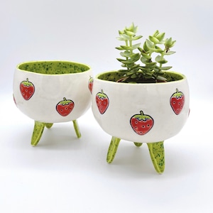 Puede incluir: Dos macetas de cerámica blanca con bordes y patas salpicados de verde. Las macetas están decoradas con fresas rojas y verdes. Una maceta tiene una pequeña planta suculenta verde en su interior.