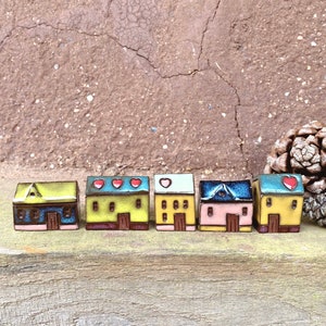 May include: Six miniature ceramic houses painted in yellow, blue and pink. Each house has a red heart on the roof and a small door. The houses are arranged in a row on a wooden surface.