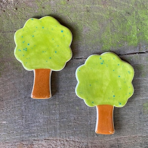 Puede incluir: Dos adornos de cerámica con forma de árbol. Las copas de los árboles son de color verde brillante con motas azules, y los troncos son marrones. Los adornos están colocados sobre una superficie de madera envejecida.