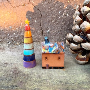 Może przedstawiać: Miniaturowy ceramiczny domek z kolorowym dachem i małym kominem, obok ceramicznej choinki w kolorach tęczy z złotą gwiazdą na szczycie. Oba stoją na drewnianej powierzchni.