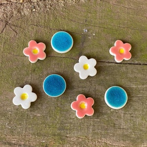 Puede incluir: Un conjunto de siete azulejos de cerámica con una variedad de diseños. Hay cuatro diseños de flores, dos en rosa, uno en blanco y uno en amarillo. También hay tres azulejos azules en forma de círculo.