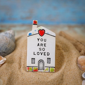 Op de afbeelding: Witte keramische huisfiguur met een rood hart en de woorden "You are so loved" aan de voorkant. Het huis heeft twee ramen en een deur. De figuur staat op een bed van zand.