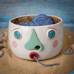 May include: A quirky, white ceramic bowl with a face design, featuring blue eyes, a green nose, and red lips. The bowl is filled with blue yarn, with a strand coming out of the mouth. The bowl is sitting on sand.