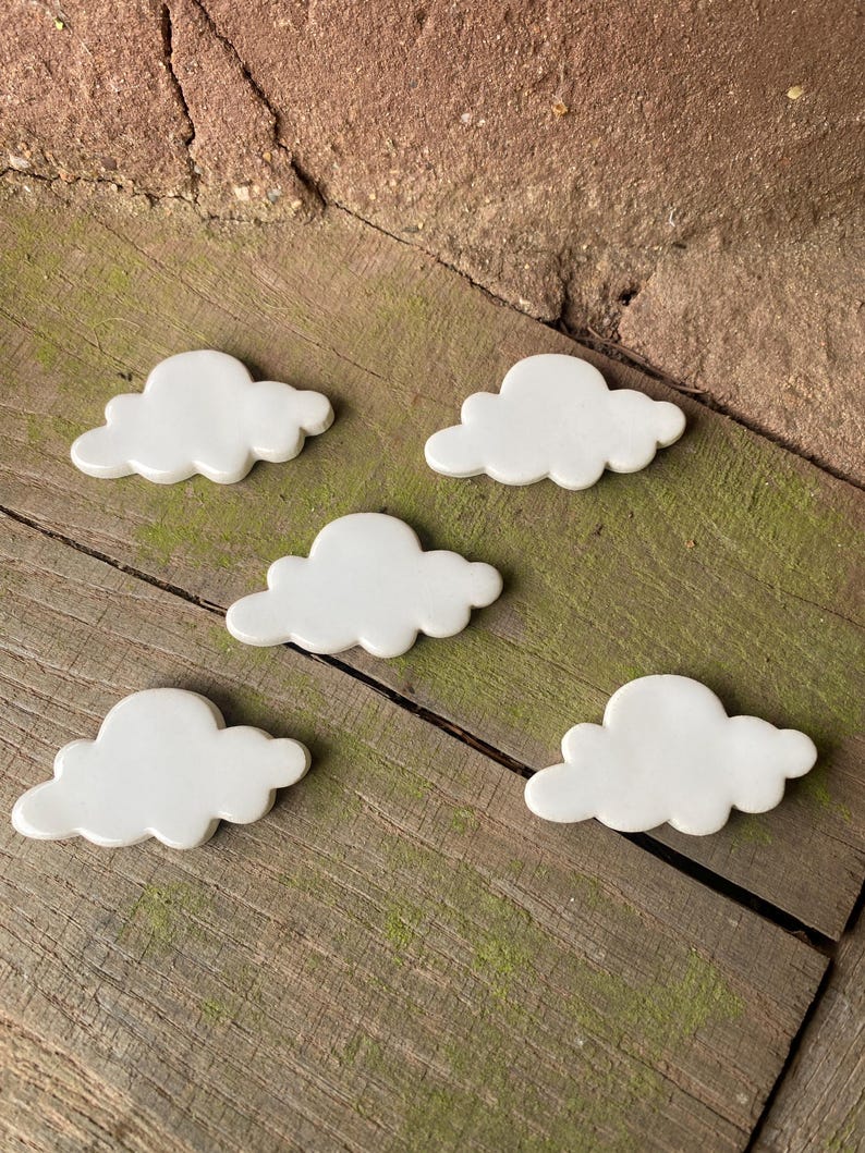 May include: Five white ceramic cloud-shaped ornaments. The clouds are arranged in a staggered pattern on a wooden surface.