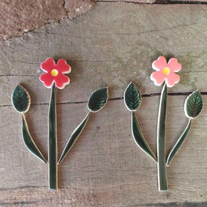 Puede incluir: Dos esculturas de flores de cerámica. Una tiene una flor roja con centro amarillo, la otra una flor rosa con centro amarillo. Ambas tienen tallos y hojas verdes. El fondo es una superficie de madera.