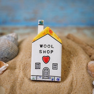 Peut inclure: Une petite maison en céramique blanche avec un toit jaune et un cœur rouge sur la façade. La maison a une cheminée et deux fenêtres. Les mots "WOOL SHOP" sont écrits sur la façade de la maison.