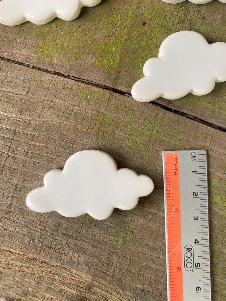 May include: White ceramic cloud shaped tiles, measuring approximately 6 cm in length. The tiles are arranged on a wooden surface with green moss.