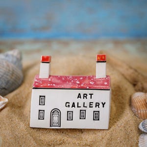 May include: A miniature white ceramic building with a red roof and the words "Art Gallery" on the front. The building is sitting on a bed of sand.