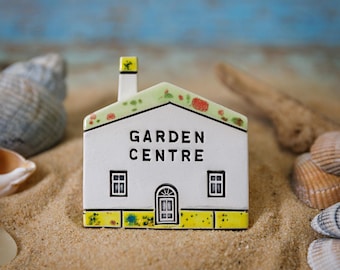 以下が含まれることがあります： 「GARDEN CENTRE」の文字が入った白いセラミック製の家の置物。屋根は緑色で赤いアクセントがあり、基部は黄色です。2つの窓とドアがあります。砂の上に置かれています。