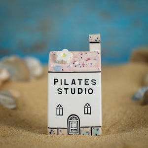 Può includere: Una casa in miniatura in ceramica bianca con tetto rosa, camino e decorazione floreale. La casa ha una porta e due finestre ad arco. Il testo "PILATES STUDIO" è stampato sul davanti.